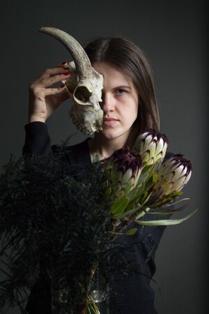 Portrait of young dark-haired girl holding half a goat skull in front of her face and a bouquet of black proteas and asparagus, duplicity conceptの写真素材