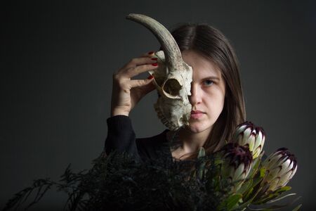 Portrait of young dark-haired girl holding half a goat skull in front of her face and a bouquet of black proteas and asparagus, duplicity conceptの写真素材