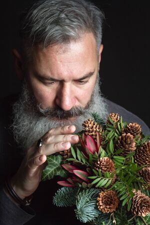 Middle-aged bearded man holding a winter bouquet of Nobilis fir twigs, cones, pistachio, leucodendron and ivy, winter gift conceptの写真素材
