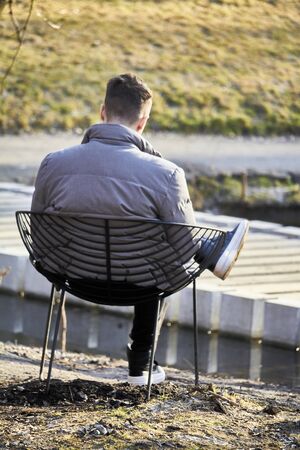 Portrait from behind of young unrecognizable young man relaxing on metal chair in city parkの写真素材
