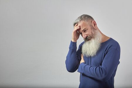 Portrait of mature gray-haired bearded man in blue sweatshirt with headache on grey background,...