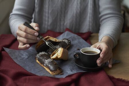 Woman eating eclairs filled with vanilla cream, traditional french eclairs with chocolate and cup of espresso, selective focus.の写真素材