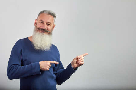 Portrait of mature gray-haired bearded man in blue sweatshirt points forefingers to side on gray backdrop, selective focusの写真素材