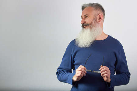 Portrait of mature gray-haired bearded man in blue sweatshirt with eyeglasses in his hand looks away on gray background, selective focusの写真素材