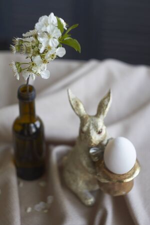 Spring composition, golden bunny figurine with egg on beige tablecloth and vase with flowering twig, Easter concept, selective focusの写真素材