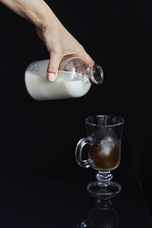 Female hand pours milk or cream from bottle into glass with ice coffee, cold coffee concept on black backdropの写真素材