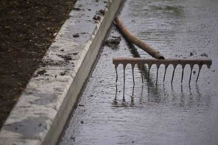 Old rusty rake lie on sidewalk near flower bed in public park on rainy day, selective focusの写真素材