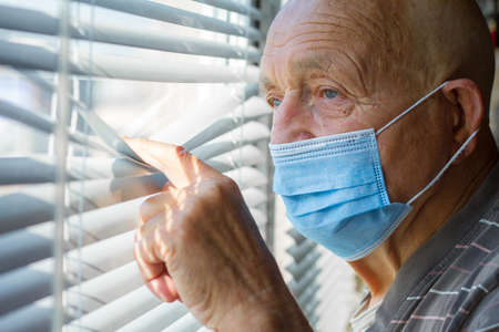 Senior citizen in blue protective mask on his face looks out through the window, stay at home conceptの写真素材