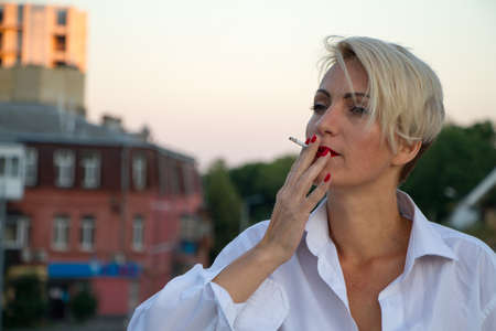 Beautiful mature blonde woman in white man shirt smokes white cigarette outdoors in city at summerの写真素材
