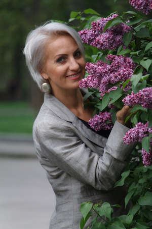 Mature blonde woman in gray suit walks in public garden, admires flowers and smiles, selective focusの写真素材
