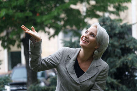 Happy mature blonde woman walks in rain and catches drops with her hand, selective focusの写真素材