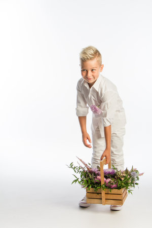 Studio portrait of fashionable blond caucasian boy with wooden basket of flowers, white backdrop, copy spaceの写真素材