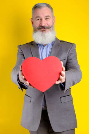 Studio portrait of mature bearded man holding out heart shape gift box, yellow background, copy spaceの写真素材