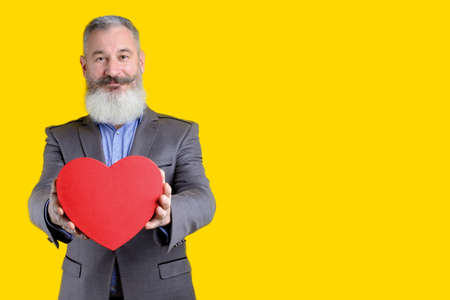 Studio portrait of mature bearded man holding out heart shape gift box, yellow background, copy spaceの写真素材