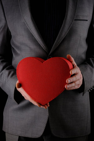 Unrecognizable man in gray suit holds out heart shaped gift red box, congratulatory concept, black backgroundの写真素材