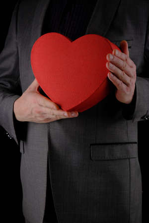 Unrecognizable man in gray suit holds out heart shaped gift red box, congratulatory concept, black backgroundの写真素材