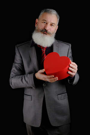 Mature bearded man wear gray suit holds red heart shaped gift box, isolated black backgroundの写真素材