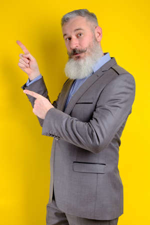 Studio portrait mature businessman dressed in gray suit points aside, I choose you this, yellow backdropの写真素材