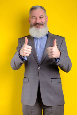 Studio portrait mature bearded businessman dressed in gray suit shows thumbs up, successful business concept, yellow backgroundの写真素材