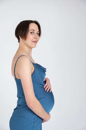 Studio portrait of young adult pregnant woman in blue dress on white grey background, happy pregnancy conceptの写真素材