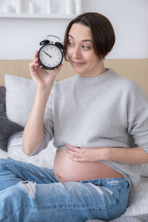 Pregnant woman with alarm clock on bed at home, giving birth soon conceptの写真素材