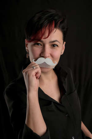 Half length portrait of young adult barber woman on black background holding metal mustache-shape comb, modern barber girl conceptの写真素材