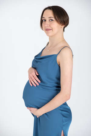 Studio portrait of young adult pregnant woman in blue dress on white grey background, happy pregnancy conceptの写真素材