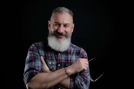 Studio portrait of mature bearded man in casual clothes with eyeglasses in hand smiles and looks...