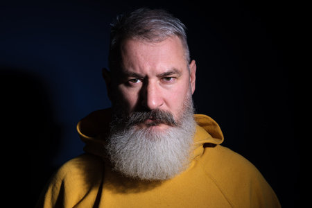 Studio portrait of mature bearded man wearing yellow hoodie looking directly at camera, blue backgroundの写真素材