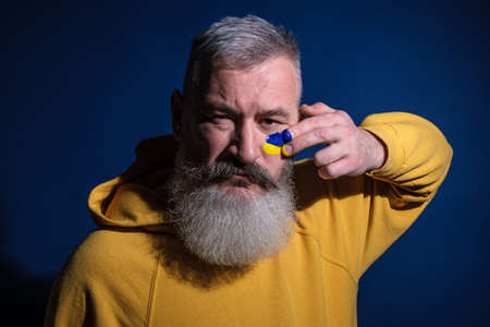 Mature bearded man dressed yellow hoodie draws Ukrainian flag on his face, protest against warの写真素材