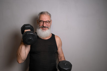 An older man with a white beard in a black tank top, holding dumbbells against a gray background. Symbolizes active aging and health at any age.の写真素材