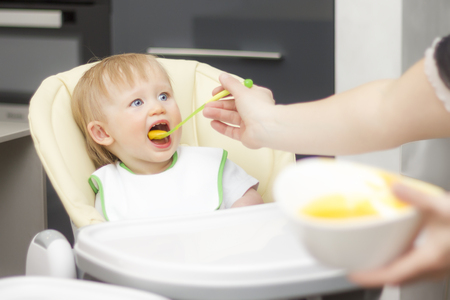 Happy toddler sitting in highchair and eating porridge. Baby learning to eat and has yogurt on porridge.の写真素材