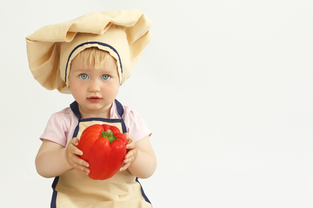 Little girl in cook clothes eats a cucumber and looks to the cameraの写真素材