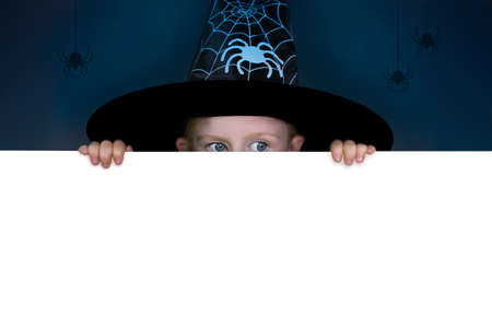 A girl wearing a witch hat peeps out from behind a white horizontal wall. Dark background and white...