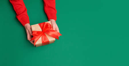 childish hands of European boy in red Christmas sweater are holding box. Gift wrapped in natural packaging and tied with red bow, on uniform green background, plenty of space, top view, copy spaceの写真素材