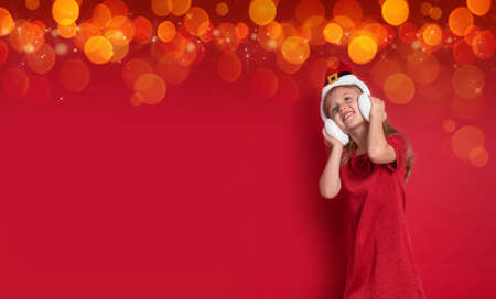 happy beautiful girl in shiny New Years dress in warm childrens headphones with Santa hat stands on a red background. Christmas banner with bokeh and flashing lights, copy space for an inscription.の写真素材