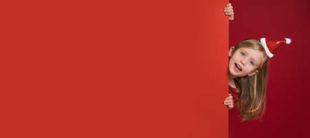 horizontal red christmas banner with copy space. A little girl in shiny Christmas clothes peeks out from behind large sheet of paper. Close-up portrait of happy child, she laughs and smiles cheerfullyの写真素材