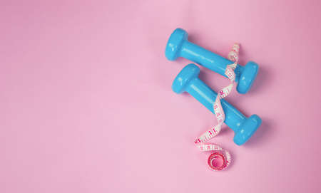 two small blue dumbbells with a tape measure close-up, side view lie on a gentle pink background with copy space. healthy lifestyle concept, body positive, female fitness, healthの写真素材