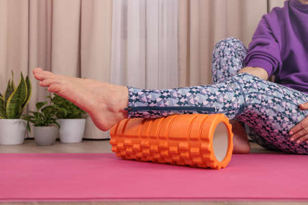 a woman in Veri Perry sportswear sits on a pink yoga mat, massages her legs with a special large, orange roll at home, improves blood flow and lymph flow, presses on acupuncture pointsの写真素材