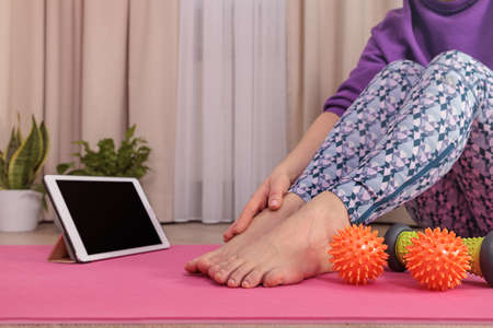 a woman in Veri Perry sportswear sits on a pink yoga mat, looks at a tablet, massages her legs at...