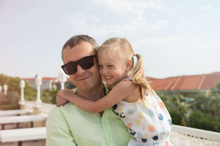 Happy father holds his daughter in his arms. European family on vacation, traveling together, on a bright sunny day. The blonde smiles, laughs and gives love to a man.の写真素材