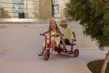 Little girl and boy ride a tricycle, play. Summer, sunny year. Hotel territory. Close up, happy childrenの写真素材