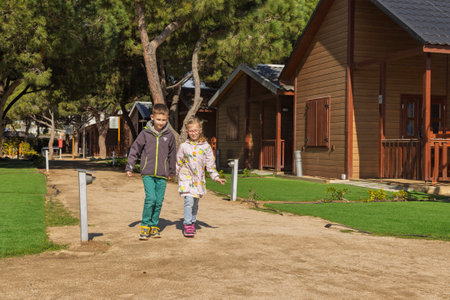Two kids run hand in hand past wooden cabins in a sunny forest. Joyful childhood, freedom, and natureの写真素材
