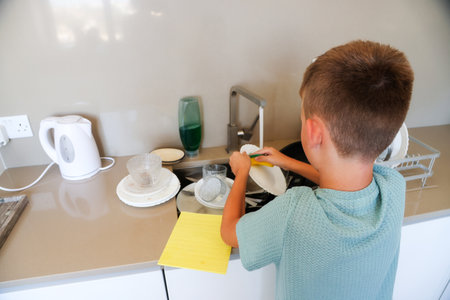 Young boy washes dish at kitchen sink, cleaning dirty plates with sponge near soap and dishes.の写真素材