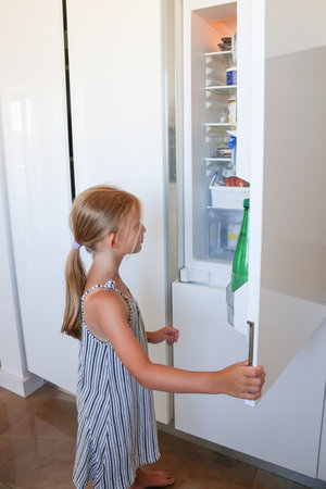 Cute child in striped dress standing barefoot and checking fridge contents in bright modern kitchenの写真素材