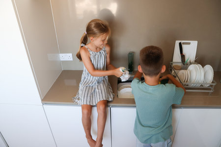 Smiling kids helping with dishwashing at home, teamwork and fun while cleaning kitchen utensilsの写真素材