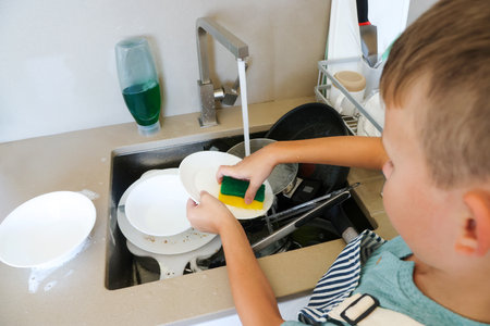 Child washes plate in sink with sponge and soap under tap water. Concept of help, chores, hygiene.の写真素材