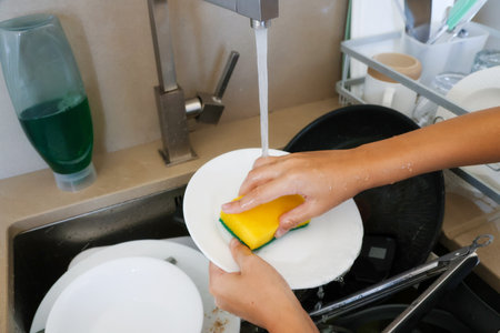 Hands clean white plate with sponge under running tap in kitchen sink, concept of home chores.