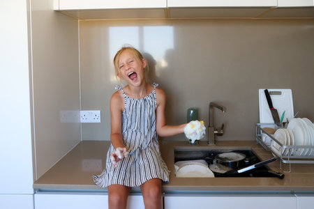 Joyful girl sitting near kitchen sink with sponge and foam, smiling, playing during dishwashingの写真素材