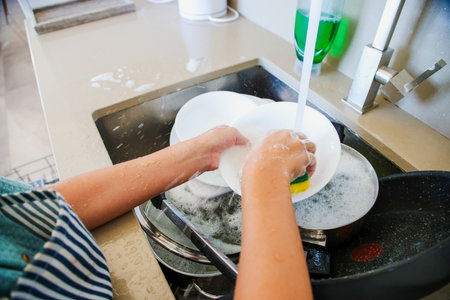 Close-up of wet hands washing white bowl with sponge in sink under water, soap and foam visibleの写真素材